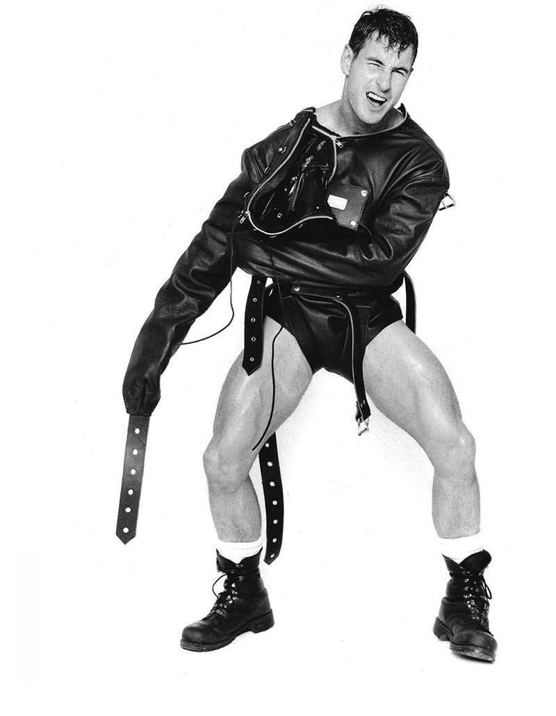 A black-and-white photo of a muscular man in a dynamic pose wearing a black leather jacket, matching leather briefs, a harness-style vest, a long leather belt with a large buckle, white socks, and black lace-up boots against a plain white background.