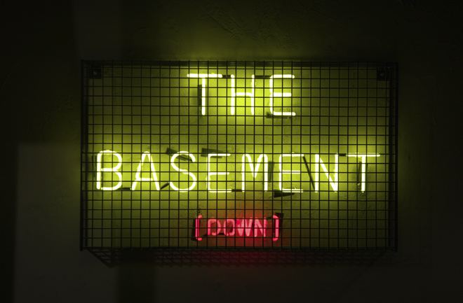 A neon sign reading THE BASEMENT, DOWN, behind a steel mesh guard