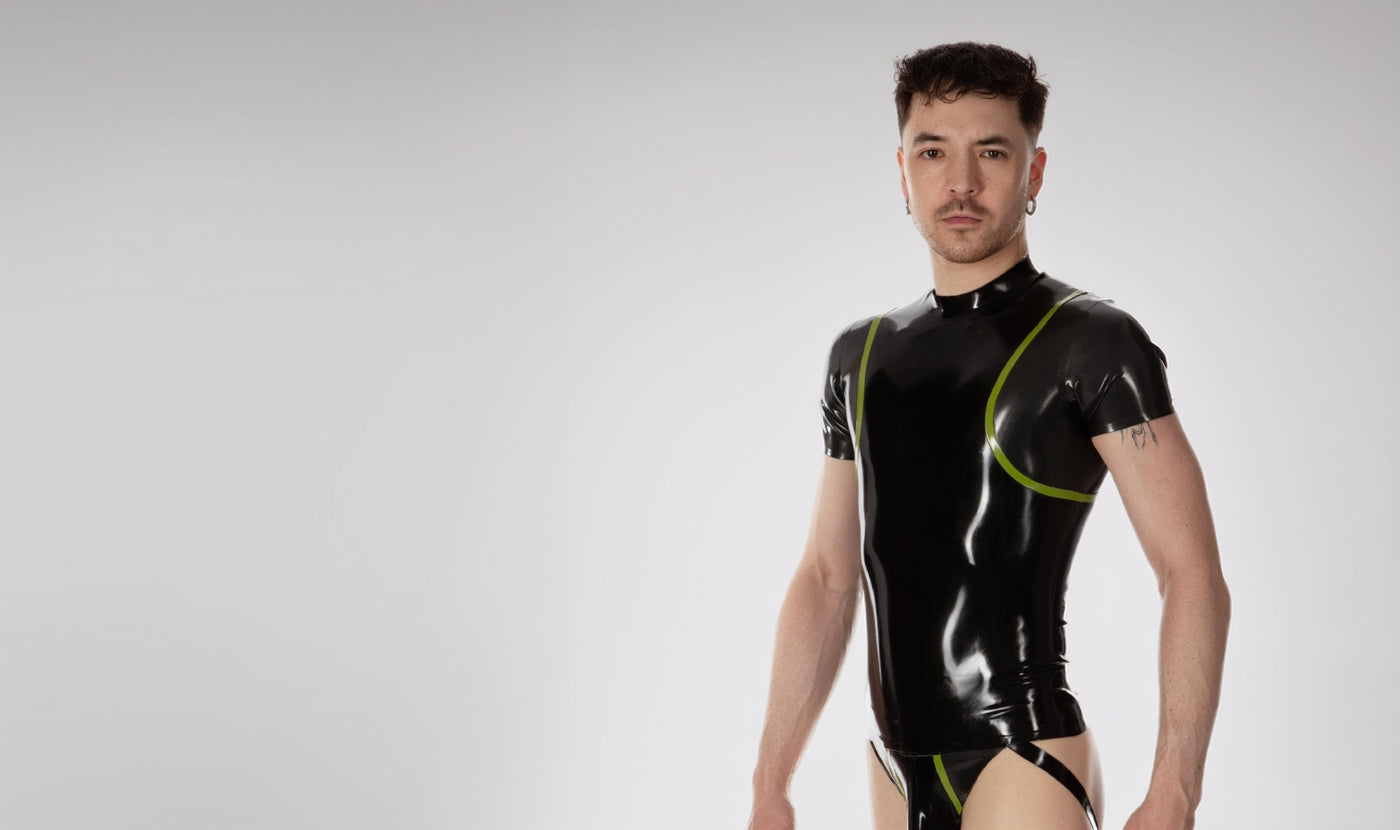 A man wearing a shiny black short-sleeve latex or latex bodysuit with lime green piping details, standing against a light gray studio background.