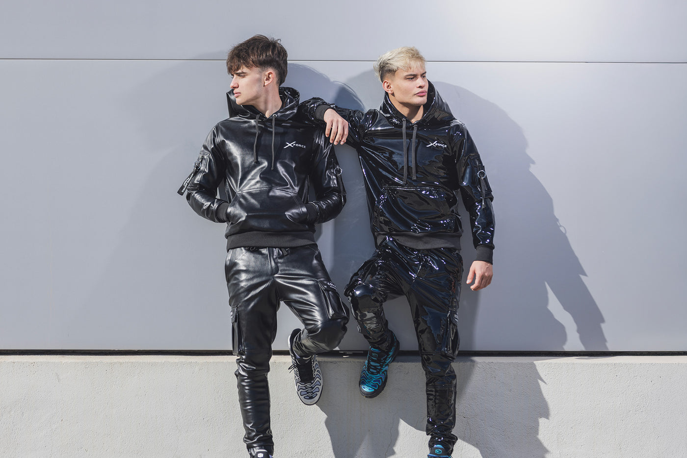 Two people wearing black reflective suits against a gray wall.